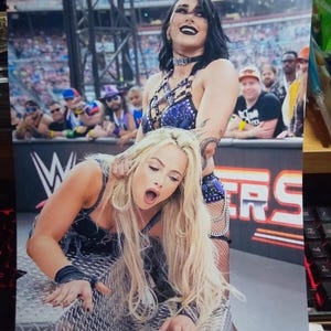 May include: A photograph of two female wrestlers in a wrestling ring. One wrestler is standing and the other is on her knees. The standing wrestler is wearing a blue and black outfit. The other wrestler is wearing a black outfit.