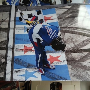 Pode incluir: Fotografia de um piloto de corridas com um fato de corrida azul e branco, inclinado sobre uma bandeira quadriculada. O piloto está numa pista pintada de azul e branco com detalhes de estrelas vermelhas. O capacete do piloto é preto.