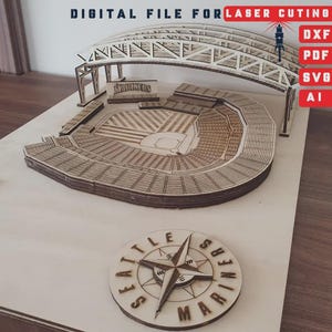 Puede incluir: Maqueta de madera cortada con láser de un estadio de béisbol, con detalles intrincados de los asientos, el campo y la estructura superior. El modelo incluye el texto "MARINERS" y una rosa de los vientos con el texto "SEATTLE MARINERS". La imagen también incluye el texto "DIGITAL FILE FOR LASER CUTTING".