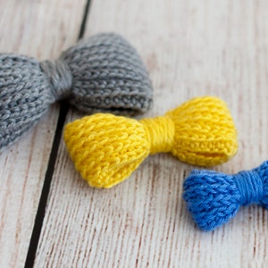 Può includere: Tre fiocchi all'uncinetto in diversi colori: grigio, giallo e blu. I fiocchi sono disposti su una superficie di legno bianco.