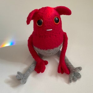 May include: A red and gray felt toy with large eyes and a small mouth. The toy has a rounded body and long arms and legs.