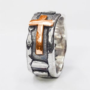 Puede incluir: Un anillo de banda de plata con una superficie texturizada y una cruz elevada hecha de cobre.