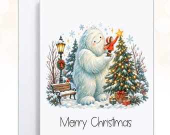 May include: A white greeting card featuring a colorful illustration of a large white yeti and a small child placing a yellow star on a decorated Christmas tree in a snowy park with a bench, a lamp post, and the text Merry Christmas below.