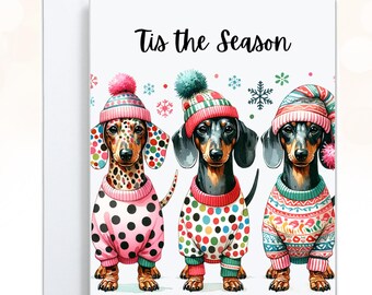 May include: A greeting card featuring three dachshunds wearing colorful winter sweaters and pom-pom hats. The dogs are decorated with polka dots and knit patterns. Above the dogs, the text reads 'Tis the Season' in black script, surrounded by small snowflakes.