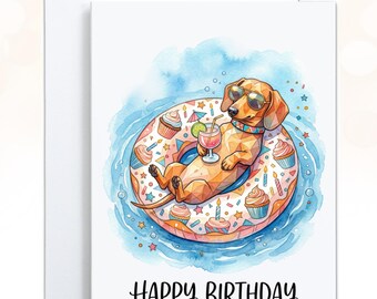 May include: A white greeting card featuring a watercolor illustration of a brown dachshund wearing sunglasses, lounging on a round pool float decorated with cupcakes and candles. The dog holds a cocktail. The text HAPPY BIRTHDAY is printed below.