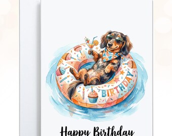 May include: A white greeting card featuring a watercolor illustration of a long-haired dachshund wearing sunglasses and a patterned bandana, lounging in a pool float decorated with cupcakes and the word BIRTHDAY. The text Happy Birthday is below.