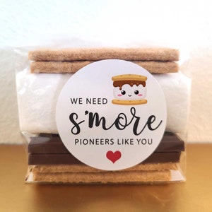 May include: A white circle sticker with the text "We need s'more pioneers like you" and a cartoon s'more with a smiling face. The sticker is on a clear plastic bag containing a s'more kit with graham crackers, chocolate, and marshmallows.