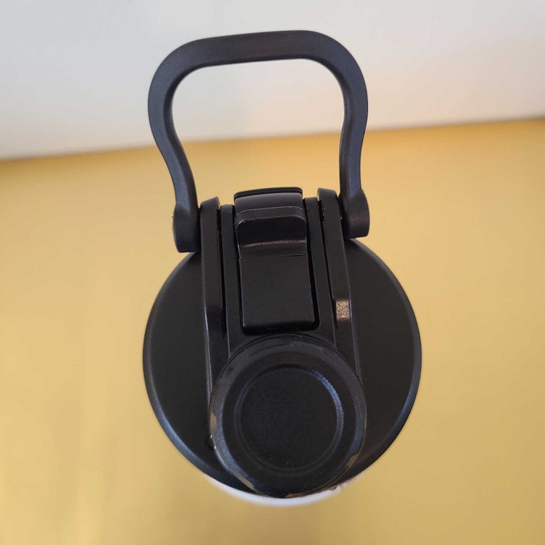 May include: A black water bottle lid with a built-in handle. The lid has a flip-top closure and a round, black cap. The handle is curved for easy carrying. The bottle is likely designed for sports or outdoor activities.