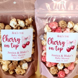 Puede incluir: Dos bolsas verticales llenas de palomitas de maíz. La bolsa izquierda es de color cobre metálico, la derecha es rosa claro. Cada una tiene una etiqueta blanca que dice: "SHE'S THE cherry on top" con una ilustración de cerezas y "Jessica & Blake's Baby Shower June 14, 2026".