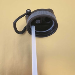 May include: A black and grey water bottle lid with a light grey straw extending downwards. The lid has a handle and a textured grip. The background is a solid gold colour.