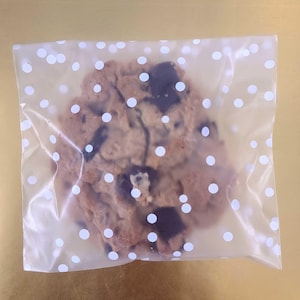 May include: A chocolate chip cookie with white chocolate chips in a clear plastic bag with white polka dots.