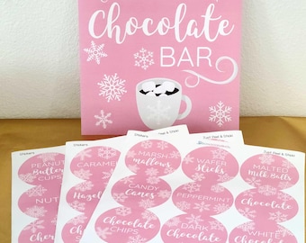 May include: A pink sign reading "Warm Up at the Hot Chocolate Bar" with snowflake and mug graphics. Below are sheets of pink stickers with white text, including "Candy Canes" and "Dark Chocolate."