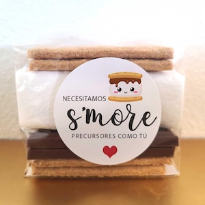 May include: A clear plastic bag filled with the ingredients for a s'more. The bag contains graham crackers, a marshmallow, and chocolate squares. A white circle sticker with a cute cartoon s'more and the text "Necesitamos s'more Precursores como tú" is on the front of the bag.