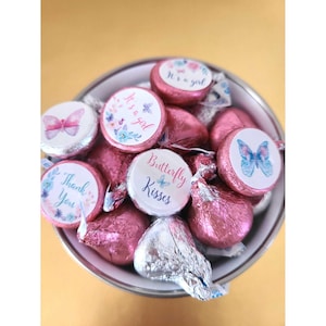 May include: A bowl filled with pink and silver foil-wrapped chocolate kisses. Each kiss has a round sticker with a butterfly design and text that says "Butterfly Kisses", "It's a girl", "Thank You", and a floral design with the text "It's a girl".