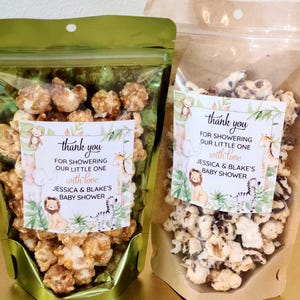 May include: Two stand-up pouches filled with popcorn. The left pouch is metallic green, the right is kraft paper. Each has a label with jungle animal illustrations and the text: thank you FOR SHOWERING OUR LITTLE ONE with love JESSICA & BLAKE'S BABY SHOWER.