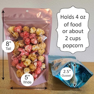 May include: Two stand-up pouches for food storage. The pouch on the left is pink and measures 8 inches tall and 5 inches wide. It is filled with popcorn and has the text "Holds 4 oz of food or about 2 cups popcorn". The pouch on the right is blue and has a 2.5 inch gusset.