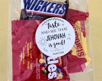 May include: A clear cellophane bag filled with assorted candies. The bag contains a Snickers bar, a red Skittles package, and a yellow Starburst package. A white circular sticker with the text "Taste and See That Jehovah is good!" is on the front.