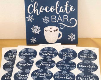 May include: A navy blue sign with white text reading "Warm Up at the Hot Chocolate Bar" with snowflake and mug illustrations. Below are oval stickers with hot chocolate toppings like marshmallows and chocolate chips.