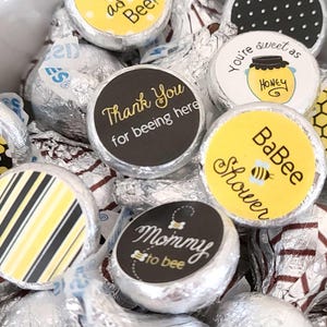 May include: Close-up of foil-wrapped chocolate candies with bee-themed labels. The labels feature phrases like "Cute as can Bee!", "Thank You for beeing here", and "Mommy to bee". The color scheme includes yellow, black, and white.