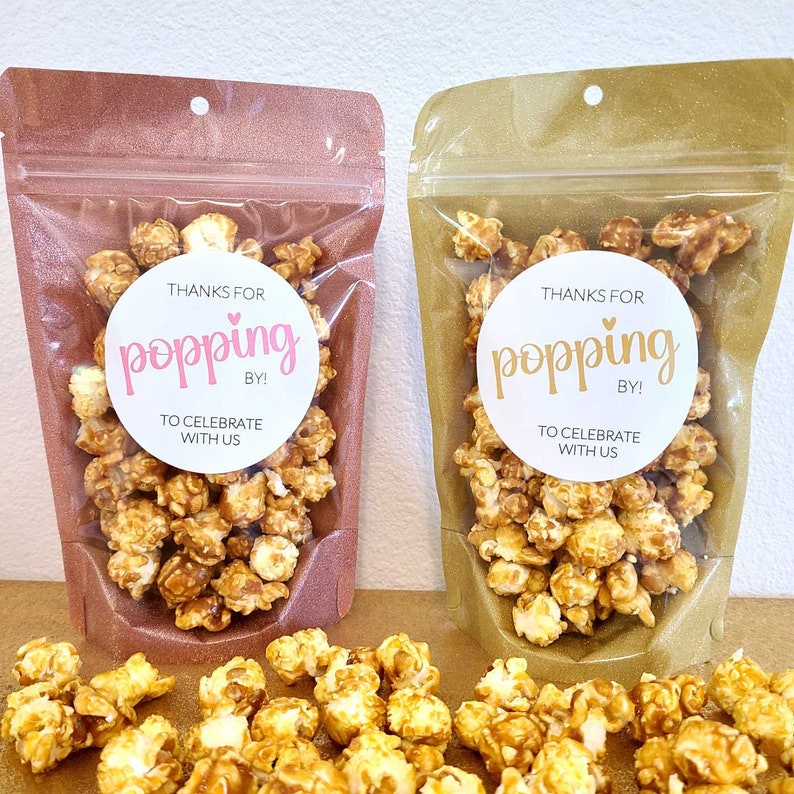 May include: Two clear plastic bags filled with caramel popcorn. Each bag has a white circle sticker with the text "Thanks for popping by! To celebrate with us." The sticker on the left bag has pink text and a pink heart. The sticker on the right bag has gold text and a white heart. The bags are sitting on a brown surface with more popcorn scattered around them.