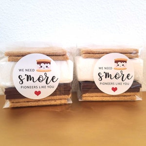 May include: Two clear plastic bags filled with s'mores ingredients. Each bag contains two graham crackers, a chocolate bar, a marshmallow, and a round sticker that says "We need s'more pioneers like you" with a cartoon marshmallow and a red heart.