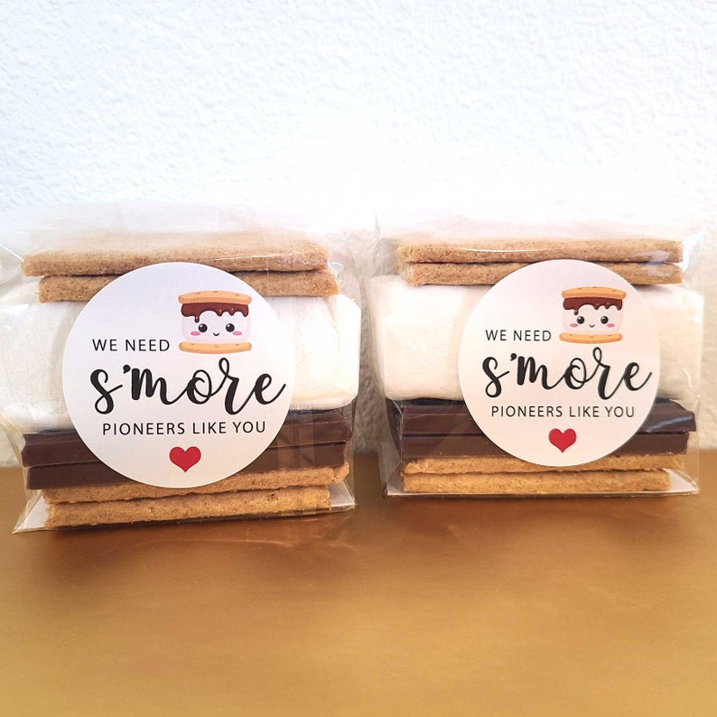 May include: Two clear plastic bags filled with s'mores ingredients. Each bag contains two graham crackers, a chocolate bar, a marshmallow, and a round sticker that says "We need s'more pioneers like you" with a cartoon marshmallow and a red heart.
