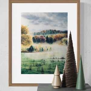 May include: A framed watercolor landscape painting depicting a misty forest scene with autumn colors. The painting is in a light brown wooden frame. Decorative wooden and fabric cone-shaped trees are on a dark gray surface.