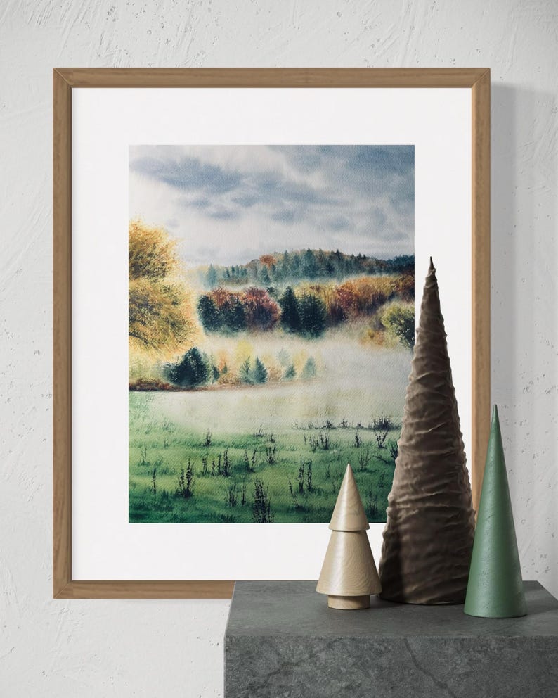 May include: A framed watercolor landscape painting depicting a misty forest scene with autumn colors. The painting is in a light brown wooden frame. Decorative wooden and fabric cone-shaped trees are on a dark gray surface.