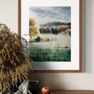 May include: A framed watercolor landscape painting depicting a misty meadow and colorful trees. The artwork is in a brown wooden frame, displayed on a wooden surface with decorative items, including a basket of dried plants and a red apple.