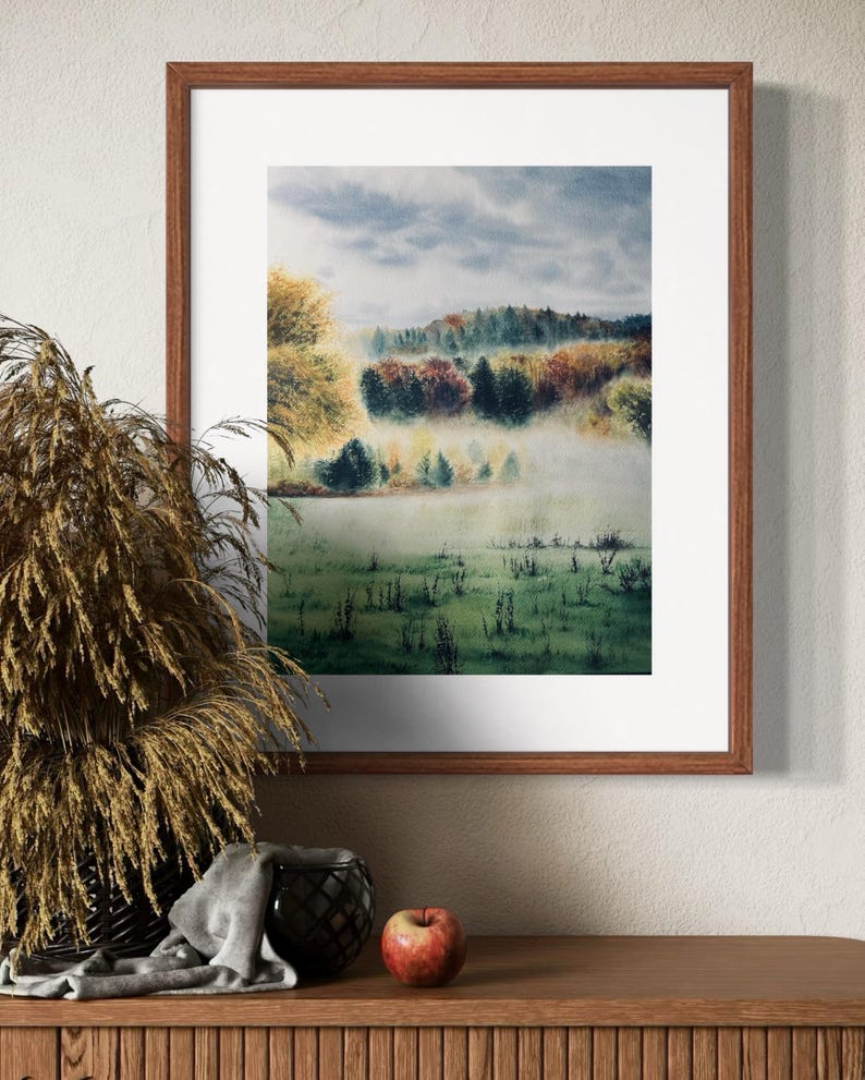 May include: A framed watercolor landscape painting depicting a misty meadow and colorful trees. The artwork is in a brown wooden frame, displayed on a wooden surface with decorative items, including a basket of dried plants and a red apple.