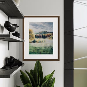 May include: A framed landscape art print featuring a misty field and colorful trees. The artwork is in a brown frame and hangs on a white wall. A dark shelf with decorative items and a plant are in the foreground.