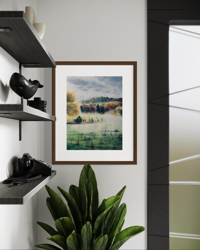 May include: A framed landscape art print featuring a misty field and colorful trees. The artwork is in a brown frame and hangs on a white wall. A dark shelf with decorative items and a plant are in the foreground.