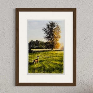 May include: Watercolor painting of a deer in a grassy field with a tree and sunset in the background. The artwork is framed in a brown wooden frame with a white mat. The painting features shades of green, yellow, and brown.