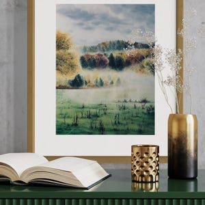 May include: A framed watercolor landscape painting with a gold frame, depicting a misty field and colorful trees. A gold vase, a gold candle holder, and an open book sit on a green cabinet.