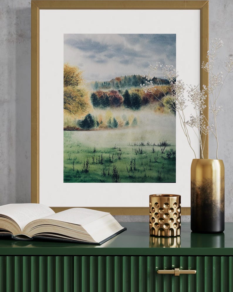 May include: A framed watercolor landscape painting with a gold frame, depicting a misty field and colorful trees. A gold vase, a gold candle holder, and an open book sit on a green cabinet.