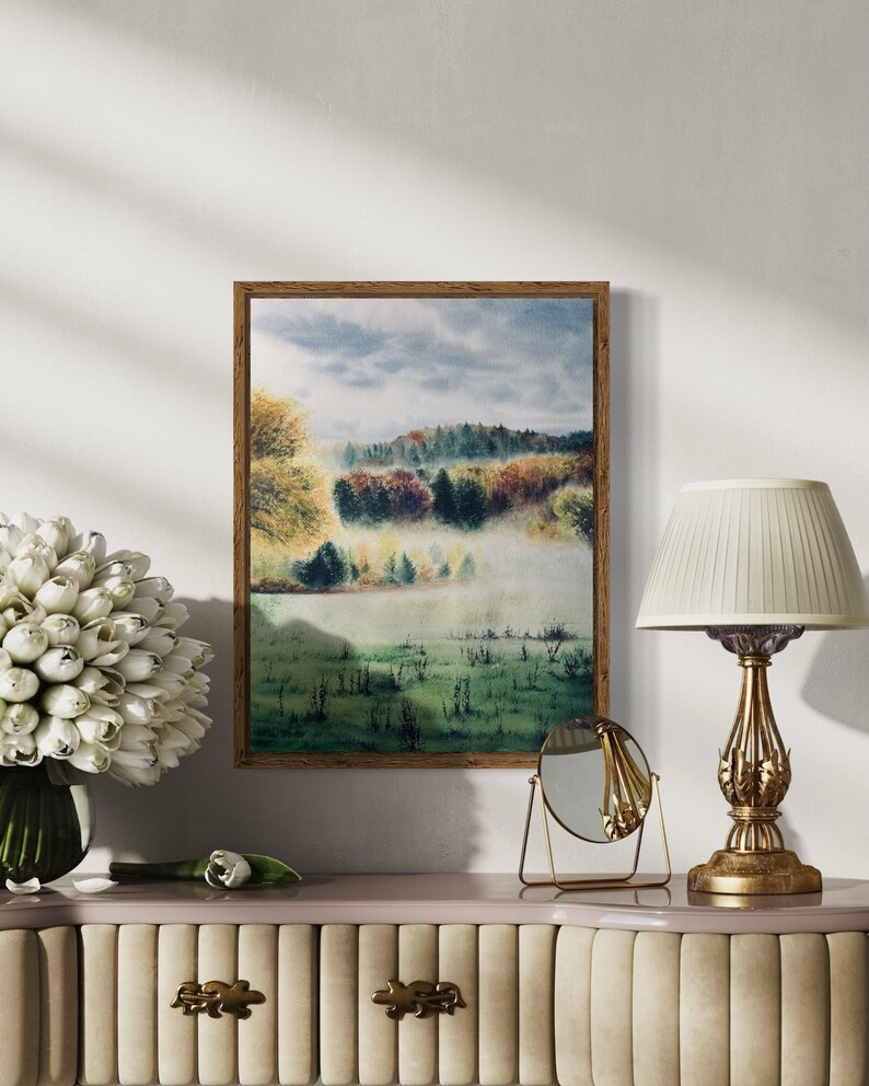 May include: A framed watercolor landscape painting depicts a misty field and trees in autumn colors. The painting is in a wooden frame and hangs above a decorative table with a lamp, mirror, and a vase of white flowers.