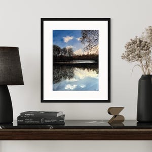 May include: A framed photograph of a lake with trees reflected in the water. The sky is blue with white clouds. The frame is black and the mat is white.