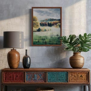 May include: A framed landscape painting depicts a misty field with trees in autumn colors. The painting hangs above a wooden console table with decorative drawers in red, teal, and gold. A lamp and vase are on the table, with a woven basket below.