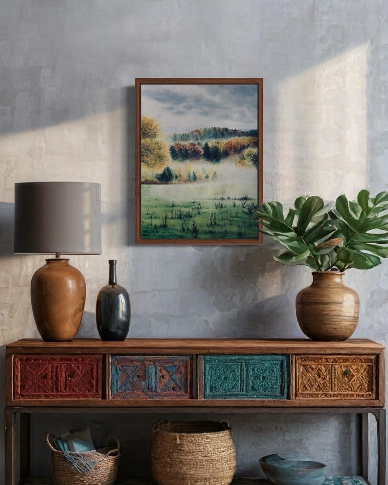May include: A framed landscape painting depicts a misty field with trees in autumn colors. The painting hangs above a wooden console table with decorative drawers in red, teal, and gold. A lamp and vase are on the table, with a woven basket below.