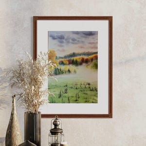 May include: A framed watercolor painting depicts a misty landscape with a field in the foreground and colorful trees in the background. The painting is in a brown wooden frame and is displayed on a wooden surface.
