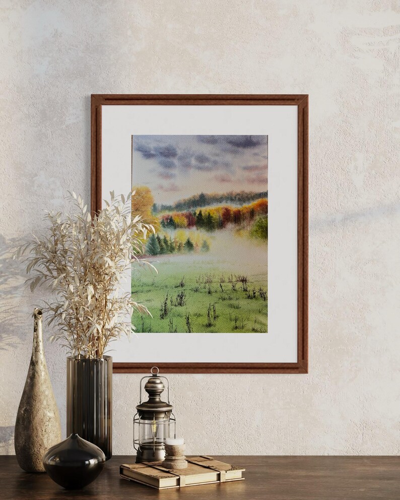 May include: A framed watercolor painting depicts a misty landscape with a field in the foreground and colorful trees in the background. The painting is in a brown wooden frame and is displayed on a wooden surface.