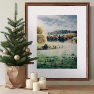 May include: A framed landscape art print with a forest scene in autumn colors. A small artificial Christmas tree with gold ornaments sits in a paper bag. White candles are on a wooden surface.
