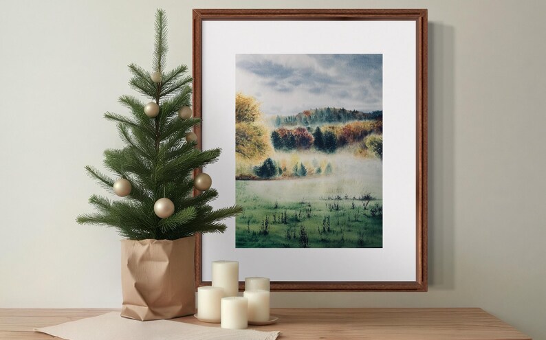 May include: A framed landscape art print with a forest scene in autumn colors. A small artificial Christmas tree with gold ornaments sits in a paper bag. White candles are on a wooden surface.