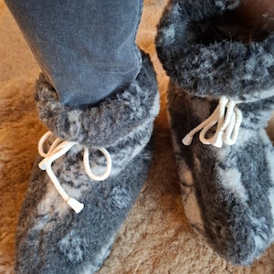 Pode incluir: Um par de pantufas de lã cinza e branca com cordões brancos. As pantufas têm uma aparência espessa e texturizada e são projetadas para cobrir o pé e o tornozelo. As pantufas são mostradas em uma superfície marrom.