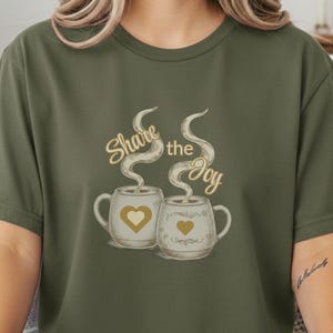Puede incluir: Camiseta verde oliva con un gráfico de dos tazas humeantes, cada una con un diseño de corazón. El texto "Share the Joy" está sobre las tazas en oro. La camiseta tiene mangas cortas.
