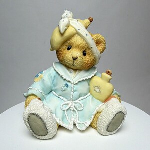 May include: A tan teddy bear figurine wearing a light blue robe with white trim and a yellow head wrap. The bear holds a yellow hot water bottle with a small white rabbit graphic on the front. The bear has textured fur and grey paw pads.
