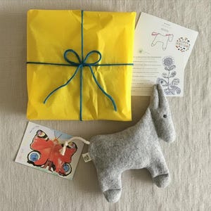 Peut inclure: Un cadeau emballé dans du papier de soie jaune vif noué avec une ficelle bleue, un âne en feutrine grise avec une queue en corde blanche, une carte illustrée d'un papillon coloré et une carte blanche avec un dessin au trait d'un cheval.