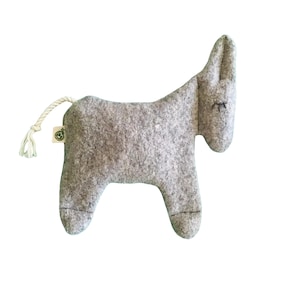 May include: A light gray felt donkey toy with a textured surface, featuring embroidered closed eyes and a white rope tail. A small square tag with a green circular logo is attached to the back near the tail, showing a simple animal silhouette.
