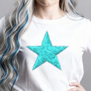 May include: White t-shirt with a large, embroidered turquoise star. The star has a textured design and a silver-coloured border. The shirt is a simple, casual style.