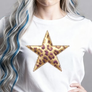 May include: White t-shirt with a large embroidered star. The star has a gold border and a leopard print pattern in gold and purple. The shirt is a casual style.